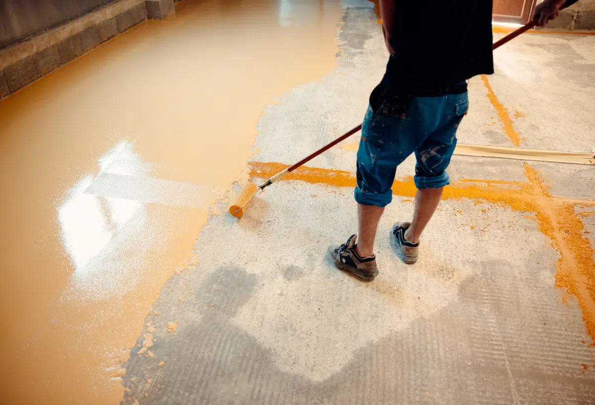Worker pouring yellow epoxy resin from a bucket onto a concrete floor
