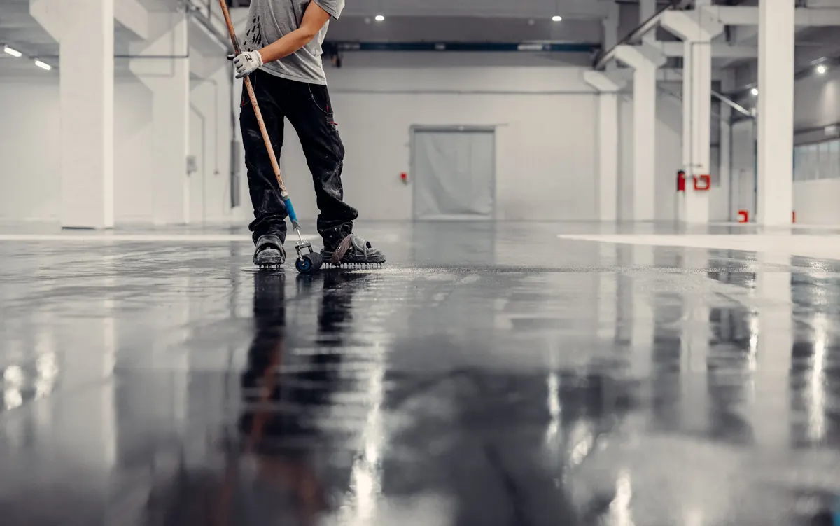 Worker using a spiked roller on a fresh dark gray epoxy floor in an industrial warehouse