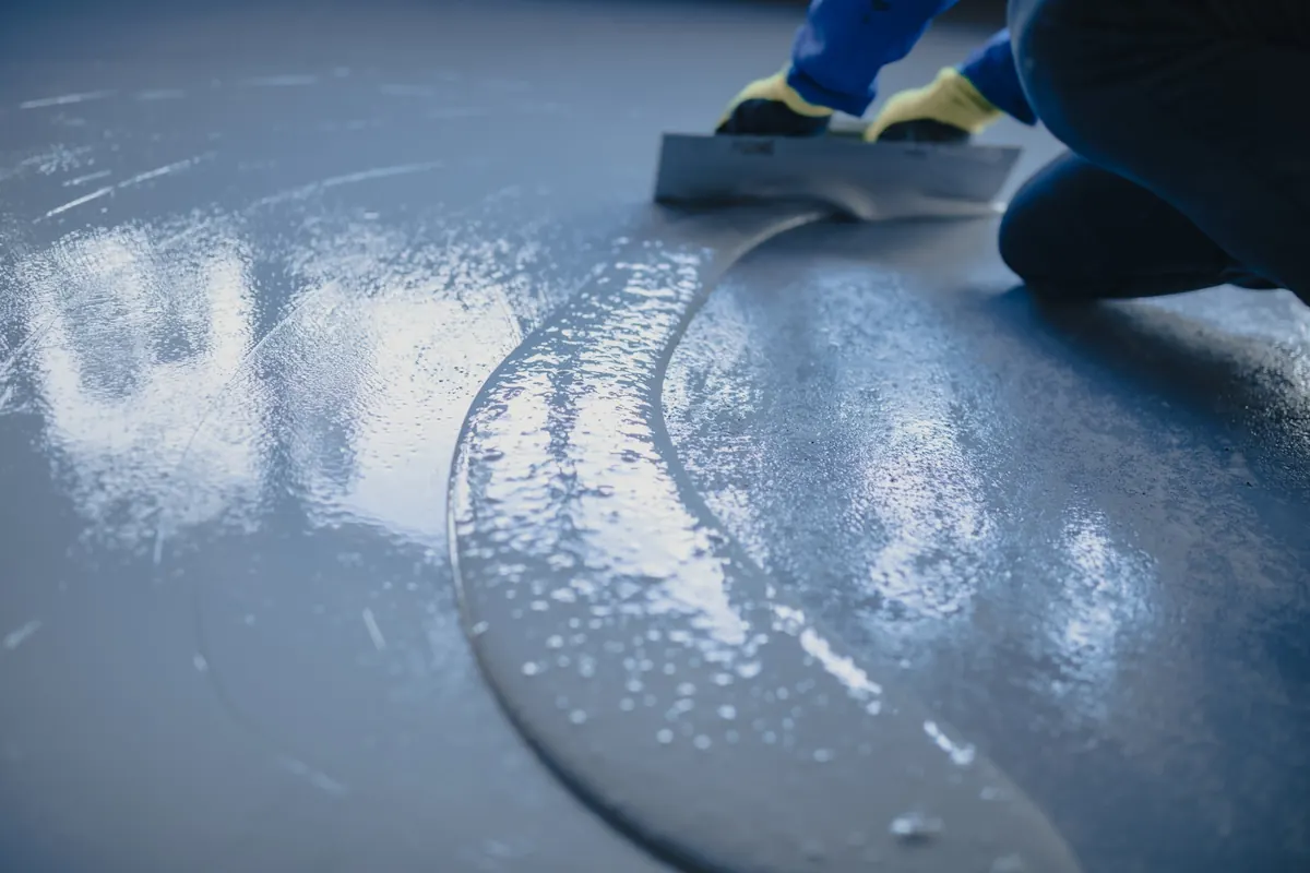 Installer smoothing a gray floor coating with a steel trowel in sweeping arcs